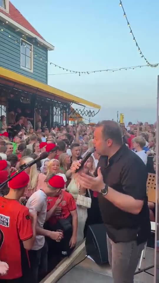 Zanger Mark Schokker zingt op een dorpsfeest in Volendam, midden tussen een straat vol publiek met rode petjes