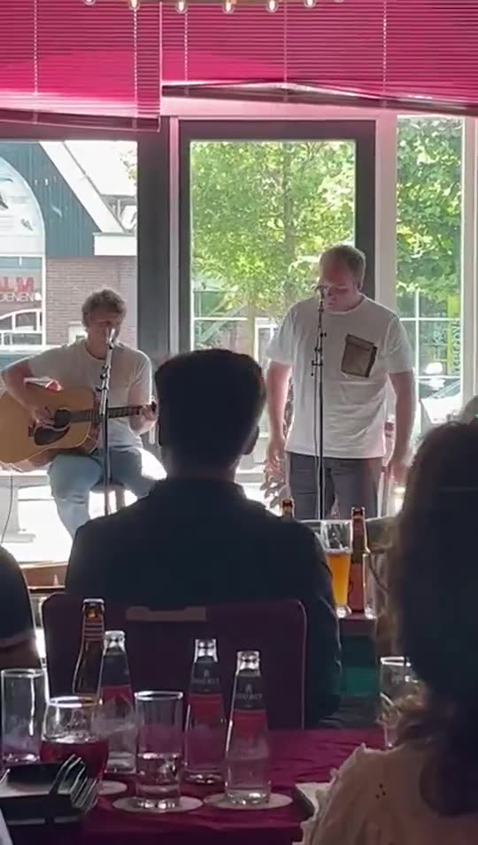 Zanger Mark Schokker zingt met een gitarist in een zaal met daglicht, met gasten aan tafel