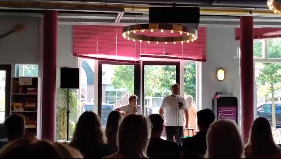 Zanger Mark Schokker zingt staand voor de ramen van een feestzaal met zittend publiek